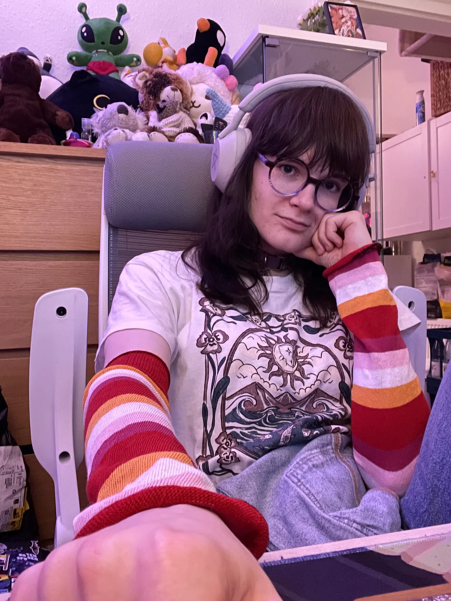Photograph. Flori sitting in a white/grey desk chair in a small, warmly lit room. Behind her is a shelf with many different plushies on it, including a kurzgesagt duck and an ikea aftonsparv. Ava looking into the camera. She is holding her own head with the left arm, and is reaching towards the camera with the other arm, presumably to take the picture. They are wearing a white tshirt with print on it, some light grey headphones, a purple collar, armwarmers in lesbian striped colors, and glasses.
