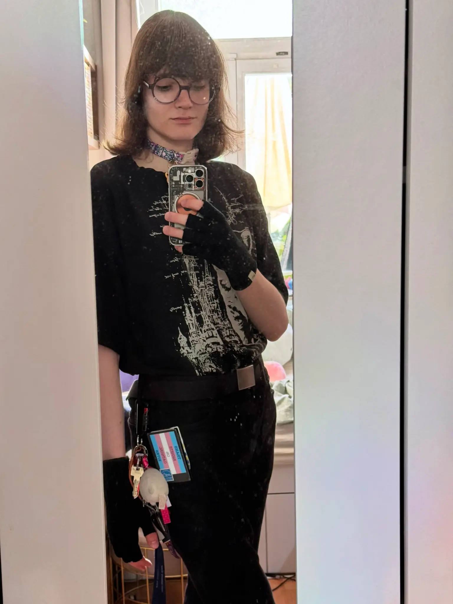 Photo, mirror selfie showing Ava with brown hair, bangs, and round glasses holding a smartphone. Flori wears a black t-shirt with a white, partially obscured graphic, black fingerless gloves, a patterned collar, and black pants with a black belt with silver clasp, and a transgender flag-bestickered wallet displaying mirrored text TRANS RIGHT OR CATGIRL BITES. A carabiner is attached to one of the belt loops of the pants, and many keys and a plushie are attached to the carabiner.