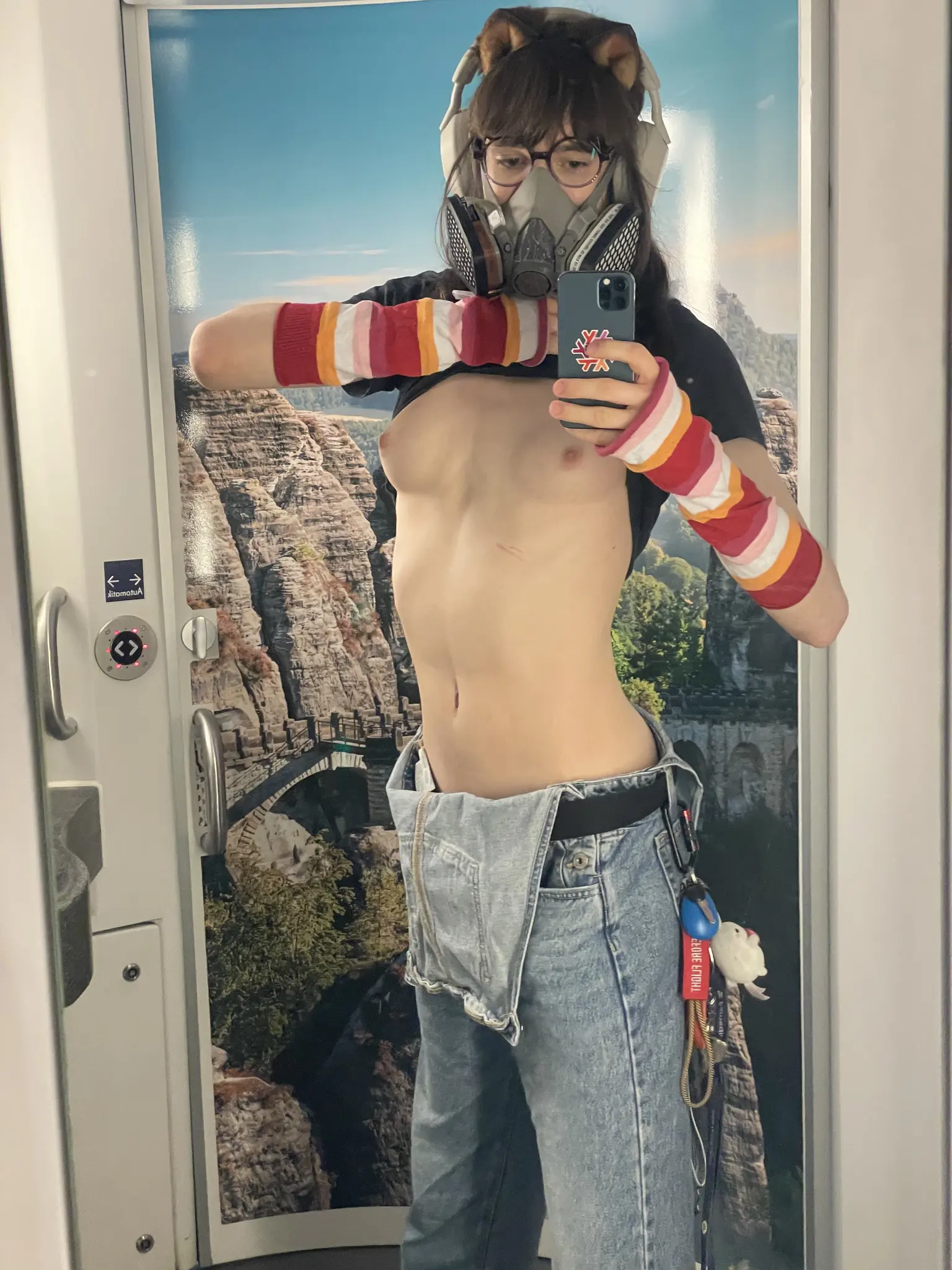 Mirror selfie of Ava in a train toilet. The door of the train toilet is in the background and has a big print of a mountain range on it. Ava pulled up her shirt to expose her nude breasts and tummy. She is wearing dungarees, the attachers of which are unhooked. On one of the belt loops, she attached a keychain using a carabiner. The keychain has a dog clicker, a small plushie and some other things dangling from it. Ava is also wearing lesbian-striped arm warmers/sleeves, white over ear headphones, cat ears which match her long brown hair very well, and a 3M-Style 6000 series half-face respirator.