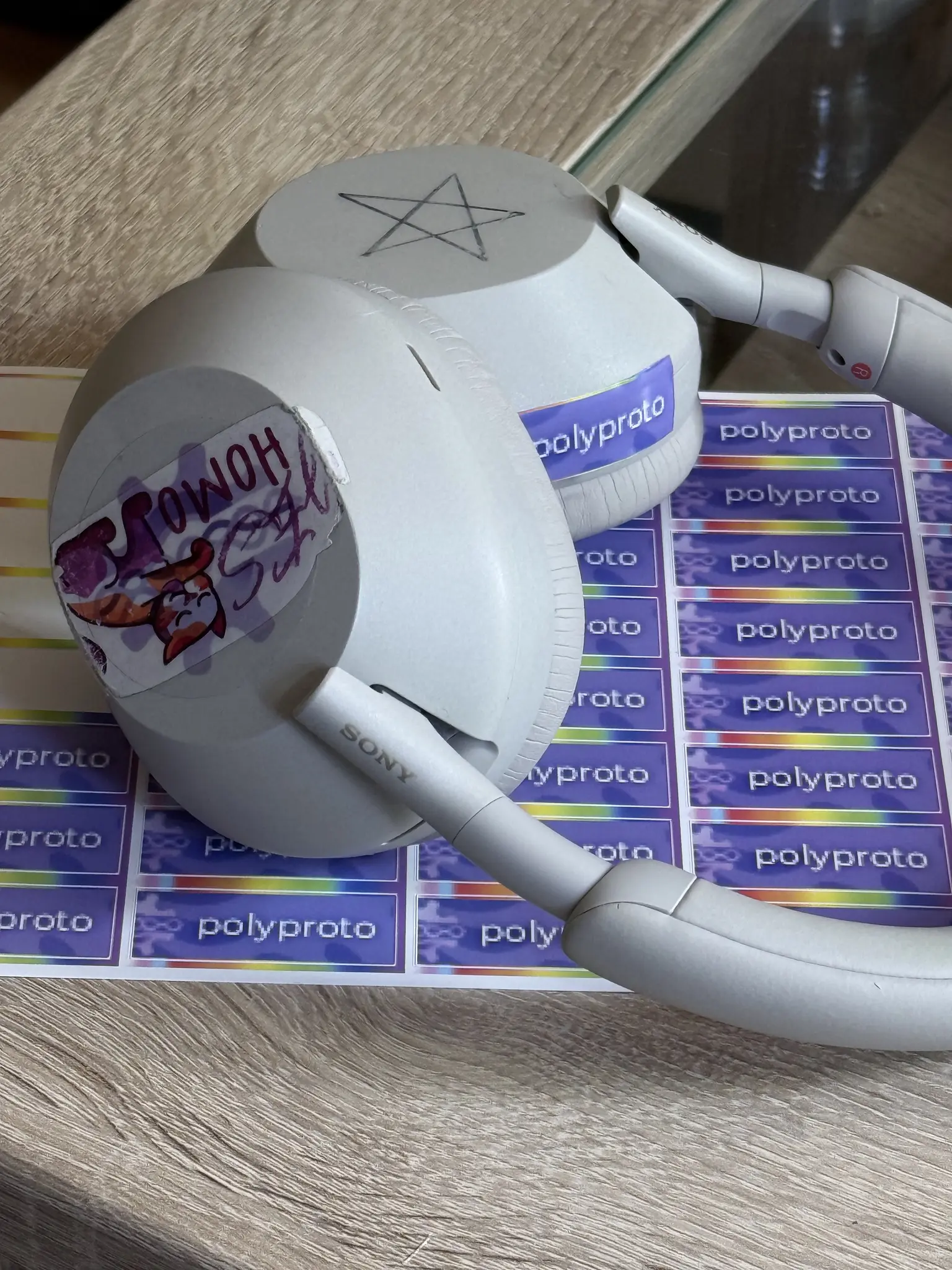 Photo of white Sony wireless headphones resting on a sheet of purple "polyproto" stickers. A pentagram is drawn on one earcup, and a sticker with a lesbian colored cat cleaning one of its front legs with its tongue and the phrase "Yes Homo" is stuck to the other earcup, with a transparent polyproto gear sticker layered above.