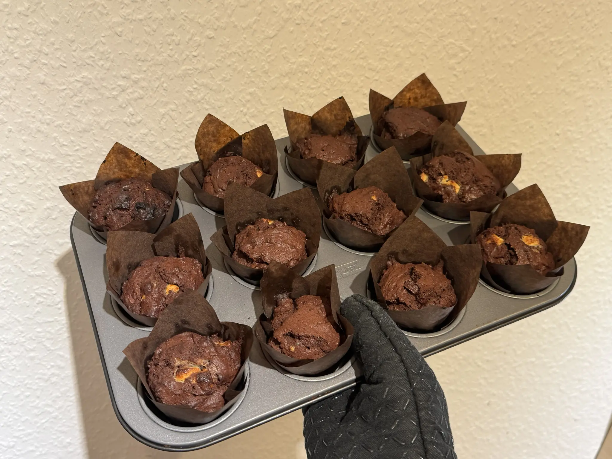 12 freshly baked triple choc muffins in brown paper liners sitting in a muffin baking mold