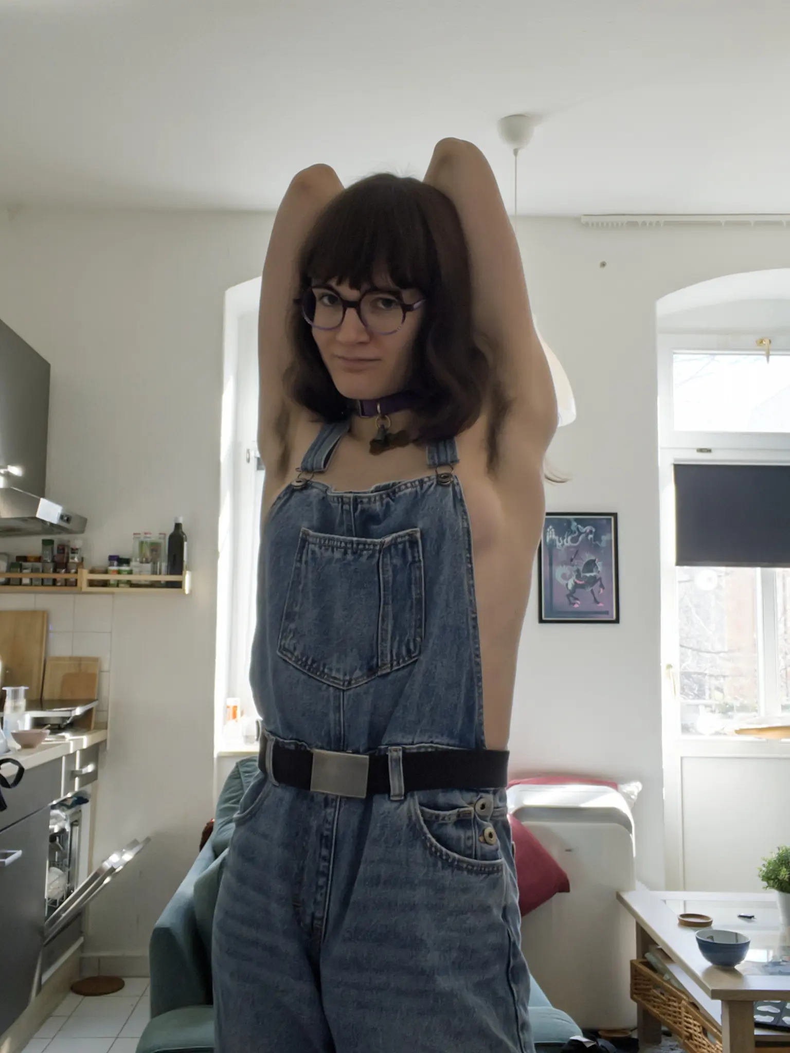 Flori in a living room/kitchen room lit by daylight. Flori is wearing a pair of dungarees and nothing else. Her boobs is peeking out a little on the side of the pouch. Both of their arms are lifted up and folded behind their back, revealing short armpit hair. Ava is smiling towards the camera. Her hair is straight, and dark brown. She is wearing a purple collar with a golden bone pendant